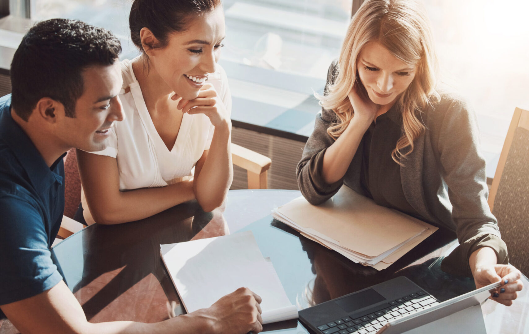 Three people sitting at a table reviewing documents and looking at a laptop, engaged in discussion.