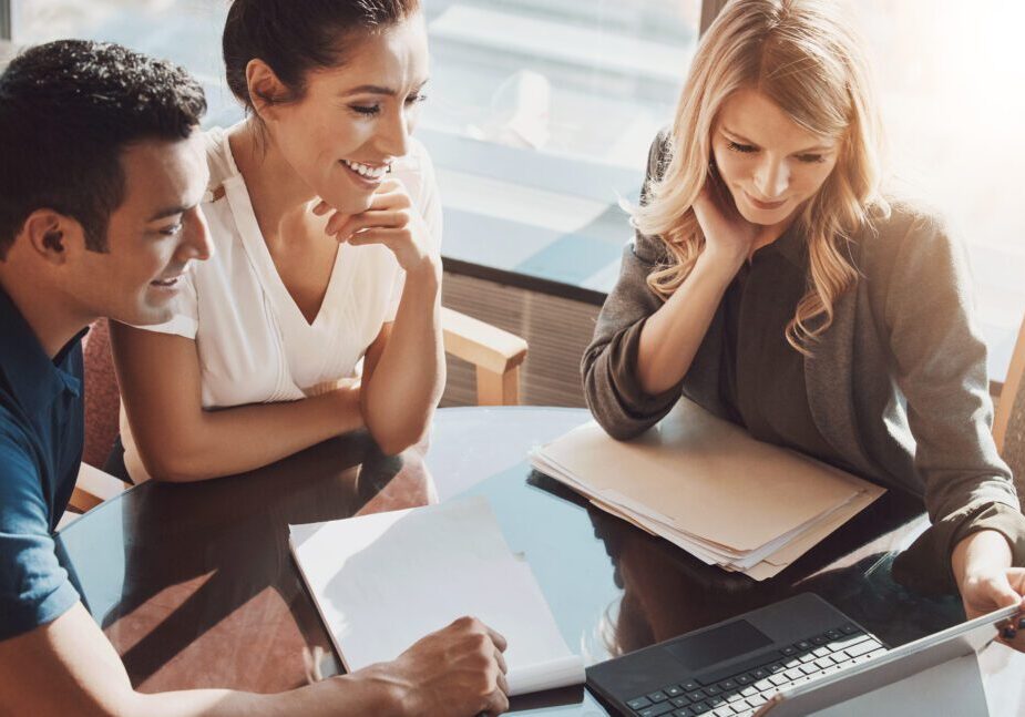 Three people sitting at a table reviewing documents and looking at a laptop, engaged in discussion.